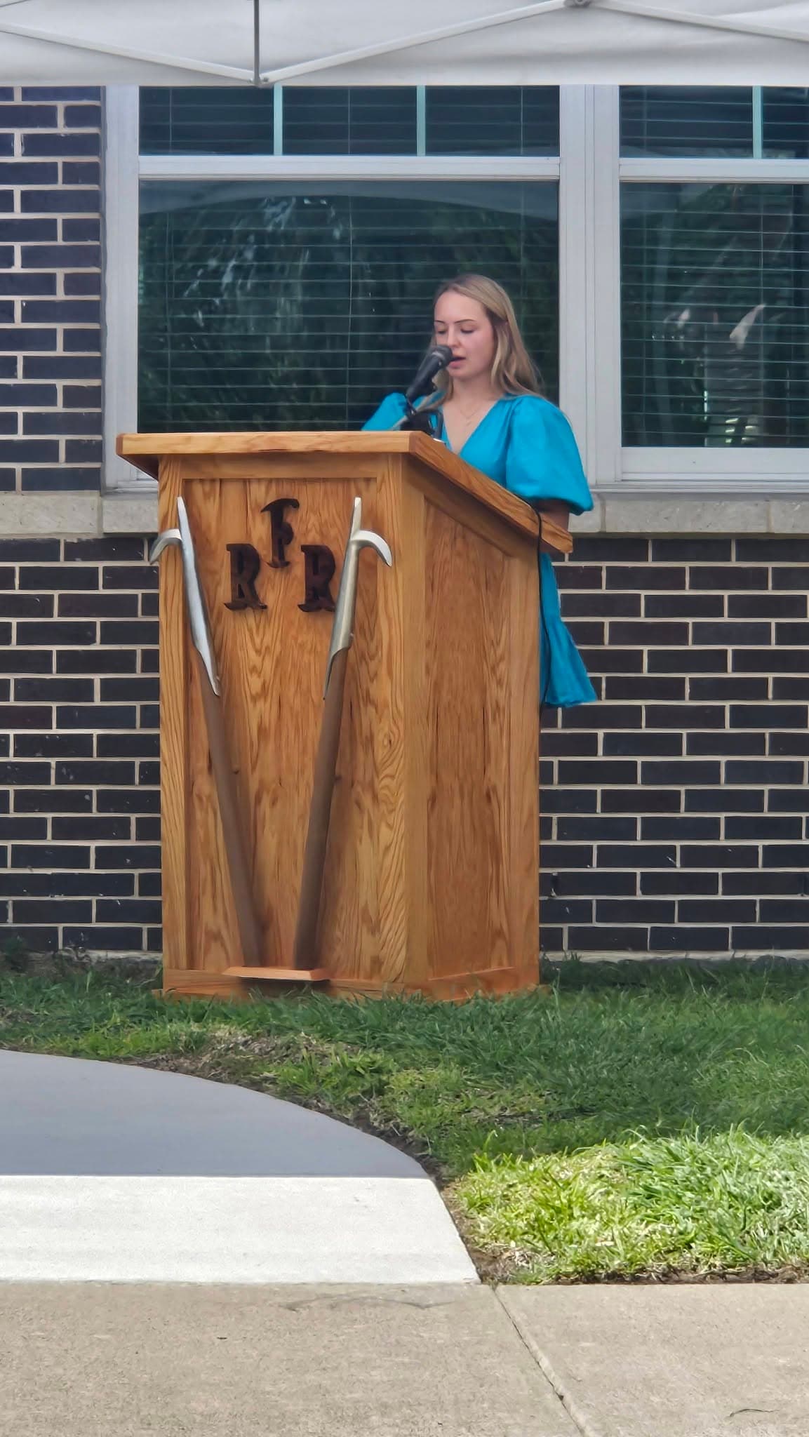 A young woman speaking at a wooden Rowlett Fire Rescue podium during the Safe Haven Baby Box dedication ceremony